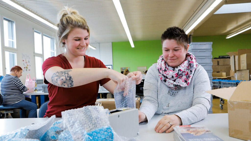 Arbeitsagogin erklärt IV-Mitarbeitenden den Arbeitsablauf beim Ausrüsten