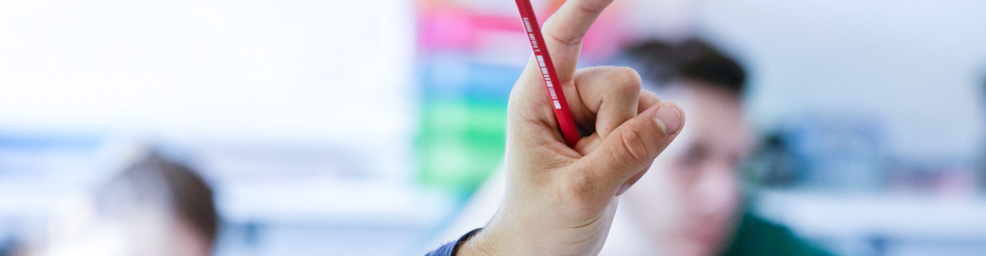 Hand mit ausgestrecktem Zeigefinger und Bleistift in einem Klassenzimmer